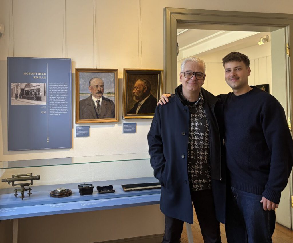 Frank Ammenn und Onno Hinne vor historischen Porträts, optischen Instrumenten und der Ausstellungstafel Hofoptiker Krille im Museum Schwerin.